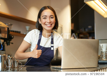 Portrait of asian girl in apron, barista in coffee shop, shows thumbs up, approve or recommend smth, working in cafe, smiling happy 97196584