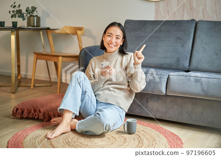 Smiling asian girl sits on floor in stylish living room, pointing finger at advertisement, showing promo banner, holding mobile phone in hand 97196605