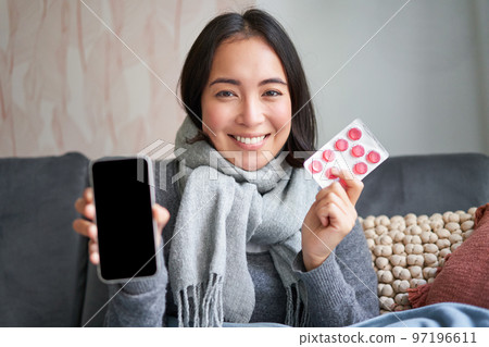 Happy asian woman catching cold, showing smartphone screen and pills, recommending online doctor app, GP or medical clinic on phone, holding drugs, staying at home with flu Happy asian woman catching cold, showing smartphone screen and pills, recommending online doctor app, GP or medical clinic on phone, holding drugs, staying at home with flu 97196611