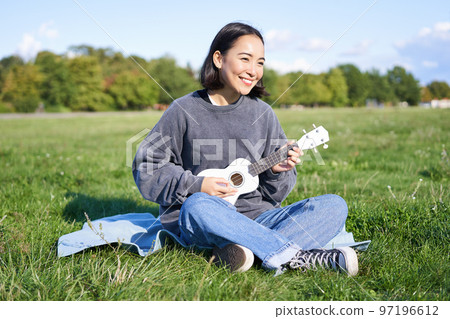 Beautiful asian girl sitting in park, playing ukulele and singing, relaxing outdoors on sunny spring day Beautiful asian girl sitting in park, playing ukulele and singing, relaxing outdoors on sunny spring day 97196612