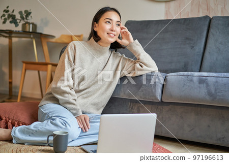 Image of smiling asian woman drinking hot tea, holding cup and sitting near laptop on floor, resting at home Image of smiling asian woman drinking hot tea, holding cup and sitting near laptop on floor, resting at home 97196613