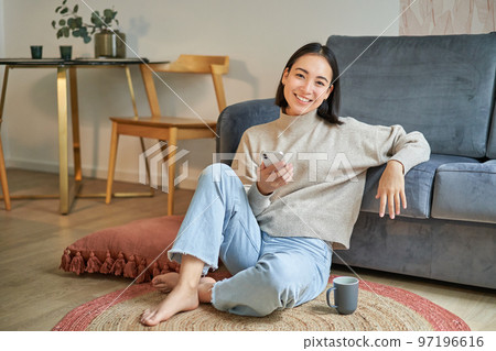 Portrait of stylish korean woman sits on floor with smartphone, using mobile phone, smiling pleased, concept of staying at home and relaxation Portrait of stylish korean woman sits on floor with smartphone, using mobile phone, smiling pleased, concept of staying at home and relaxation 97196616
