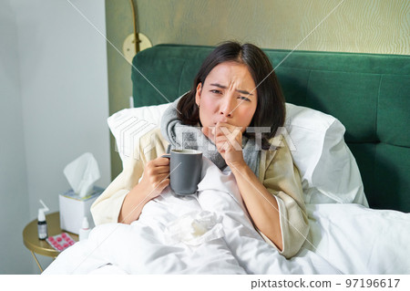 Portrait of couching asian girl in bed, feeling sick, catching cold and staying at home, looking unhealthy, drinking hot tea 97196617
