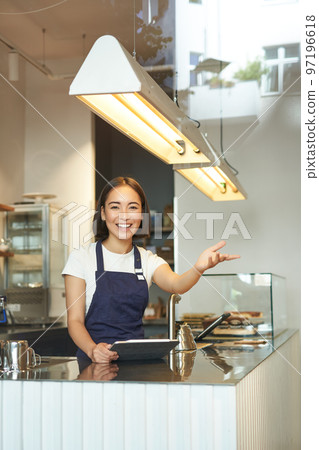 Happy asian girl barista works in coffee shop, waves at client, processing orders behind counter in cafe, wears blue apron Happy asian girl barista works in coffee shop, waves at client, processing orders behind counter in cafe, wears blue apron 97196618