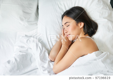 Top view image of asian woman sleeping alone in king size bed on white pillows. Young girl lying in her bedroom with eyes closed, morning sunlight shines in room Top view image of asian woman sleeping alone in king size bed on white pillows. Young girl lying in her bedroom with eyes closed, morning sunlight shines in room 97196619