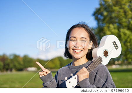 Portrait of asian smiling girl, holding ukulele over shoulder, pointing finger at copy space, banner or logo Portrait of asian smiling girl, holding ukulele over shoulder, pointing finger at copy space, banner or logo 97196620