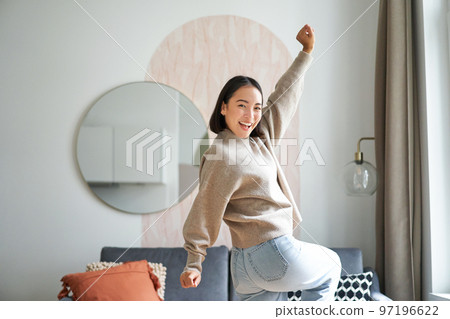 Cute excited asian woman dancing, raising hands up in triumph, celebrating and singing, standing at home in warm jamper Cute excited asian woman dancing, raising hands up in triumph, celebrating and singing, standing at home in warm jamper 97196622