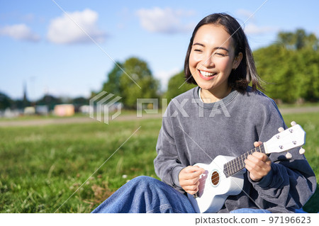 Carefree asian girl singing and playing ukulele in park, sitting on grass, musician relaxing on her free time outdoors Carefree asian girl singing and playing ukulele in park, sitting on grass, musician relaxing on her free time outdoors 97196623