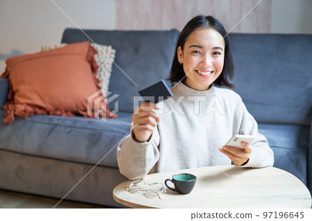 Smiling cute asian woman using credit card and smartphone, paying bills online, holding mobile phone, looking at camera 97196645