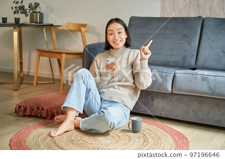 Smiling asian girl sits on floor in stylish living room, pointing finger at advertisement, showing promo banner, holding mobile phone in hand 97196646