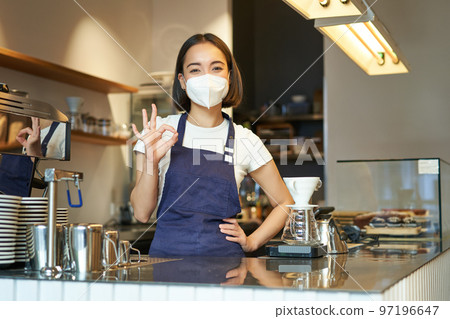 Asian female barista, girl in medical mask, shows okay sign, works in cafe behind counter, brews coffee, works with clients during covid pandemic Asian female barista, girl in medical mask, shows okay sign, works in cafe behind counter, brews coffee, works with clients during covid pandemic 97196647