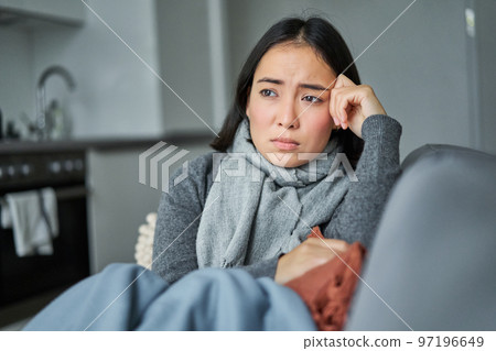Image of sad young woman feeling sick, being unwell, sitting with scarf and frowning in her living room, catching cold and staying at home on isolation Image of sad young woman feeling sick, being unwell, sitting with scarf and frowning in her living room, catching cold and staying at home on isolation 97196649