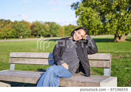 Smiling young asian woman in outdoor jacket, sitting on bench in green sunny park, resting alone, relaxing on fresh air Smiling young asian woman in outdoor jacket, sitting on bench in green sunny park, resting alone, relaxing on fresh air 97196650