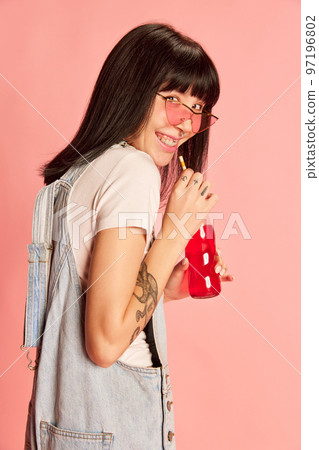Young stylish girl posing in denim overalls, smiling, drinking soda isolated on pink background. Modern style Young stylish girl posing in denim overalls, smiling, drinking soda isolated on pink background. Modern style 97196802
