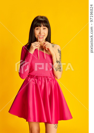 Portrait of young funny girl in bright pink dress posing with chopsticks in mouth isolated on vivid yellow background. Concept of youth, beauty, fashion, lifestyle, emotions 97196824