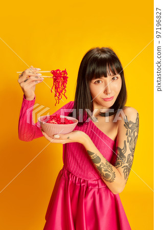 Young beautiful girl with tattoos in bright pink dress, eating pink noodles isolated on vivid yellow background. Concept of youth, beauty, fashion, lifestyle, emotions Young beautiful girl with tattoos in bright pink dress, eating pink noodles isolated on vivid yellow background. Concept of youth, beauty, fashion, lifestyle, emotions 97196827