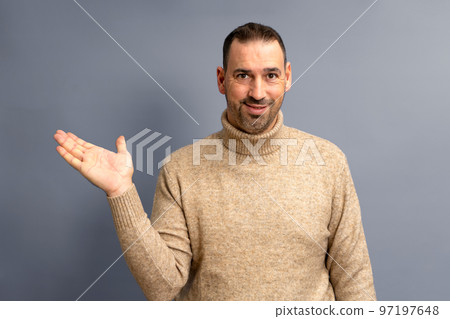 Hispanic man with beard and brown turtleneck sweater in friendly attitude offering something on the palm of his hand, isolated on gray background. Hispanic man with beard and brown turtleneck sweater in friendly attitude offering something on the palm of his hand, isolated on gray background. 97197648