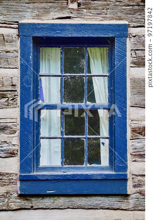 Old faded light wood building with a dark blue wooden window frame and white curtains pulled back 97197842