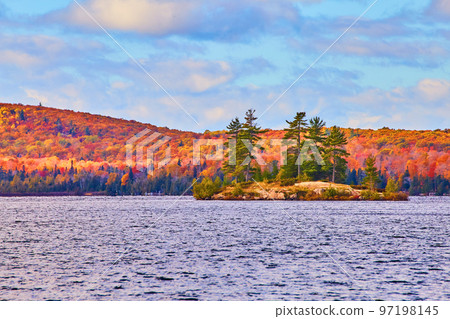 Small island of pine trees atop a rock with a fall forest behind it and a choppy lake Small island of pine trees atop a rock with a fall forest behind it and a choppy lake 97198145
