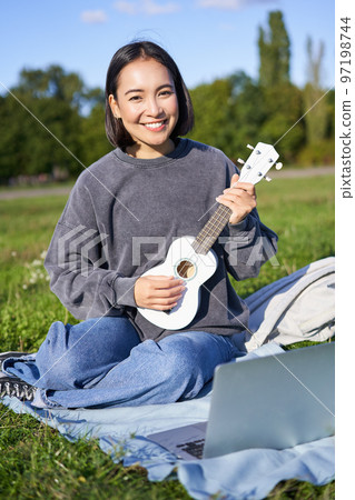 Cute korean girl, musician sits in park, plays ukulele and sings, looks up chords and tutorials on laptop 97198744