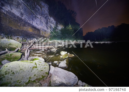 Rocky bank of a river at night with a cliff and pine trees and a dusk sky 97198748