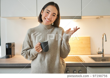 Portrait of stylish asian woman standing in kitchen with mug, drinking coffee, explaining smth, showing banner or advertisement Portrait of stylish asian woman standing in kitchen with mug, drinking coffee, explaining smth, showing banner or advertisement 97198756