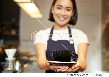 Portrait of smiling asian girl barista, waitress in cafe uniform, giving you cup of coffee, prepare drink for client, looking friendly Portrait of smiling asian girl barista, waitress in cafe uniform, giving you cup of coffee, prepare drink for client, looking friendly 97198758
