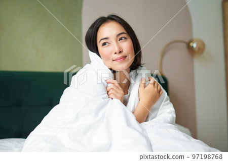 Beautiful romantic woman covers herself with warm duvet, enjoys sleeping and staying in bed, waking up happy in morning, sitting in bedroom 97198768
