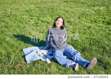 Young asian girl enjoying sunny day outdoors. Happy student having picnic on grass in park, playing ukulele Young asian girl enjoying sunny day outdoors. Happy student having picnic on grass in park, playing ukulele 97198802