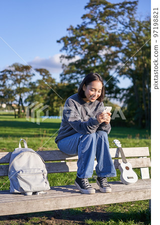 Young woman sitting in park on bench with ukulele, looking at smartphone, reading message on mobile phone and smiling 97198821