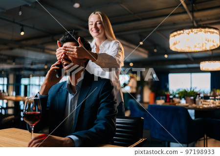 Cheerful blonde young woman closed eyes of surprised elegant man in suit sitting at table with glass red wine waiting her. Smiling beautiful female came on date with happy male. Love, dating concept. Cheerful blonde young woman closed eyes of surprised elegant man in suit sitting at table with glass red wine waiting her. Smiling beautiful female came on date with happy male. Love, dating concept. 97198933