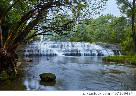 Wide shot of large cascading waterfalls with tree hanging over river and green forest 97199484