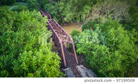 Aerial view above large metal bridge for train tracks over river surrounded by green trees Aerial view above large metal bridge for train tracks over river surrounded by green trees 97199509