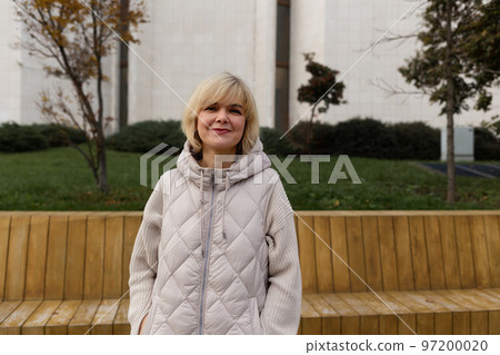 Portrait of Caucasian adult smiling blonde woman in jacket posing in city park. Concept of carefree and psychology 97200020