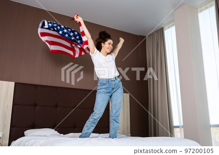 Young happy woman waving American flag on her bed. Indoor. The concept of national American holidays and patriotism. Side view 97200105