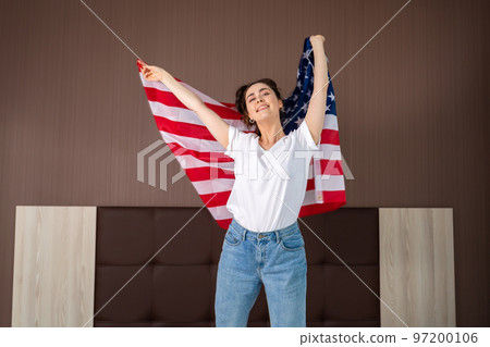 A young caucasian happy woman waves an American flag. Indoor. The concept of the American national holidays and patriotism A young caucasian happy woman waves an American flag. Indoor. The concept of the American national holidays and patriotism 97200106