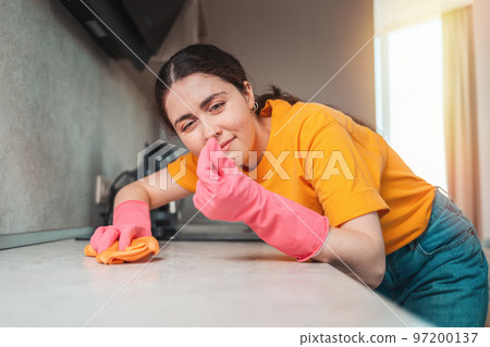 Housework. Portrait of a young woman in rubber gloves wiping the table with a rag and rubs the dirt between her fingers. Close-up portrait from the side Housework. Portrait of a young woman in rubber gloves wiping the table with a rag and rubs the dirt between her fingers. Close-up portrait from the side 97200137