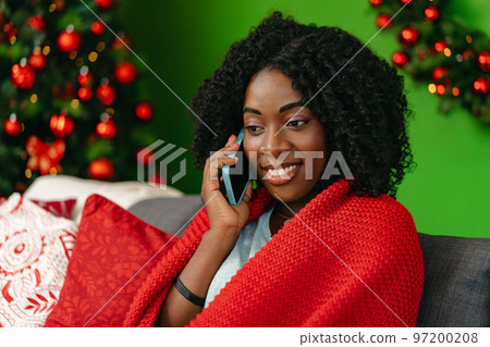 Young african woman talking on the smartphone sitting by christmas tree at home 97200208