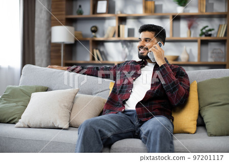 Handsome bearded positive man sitting on couch and calling to his friend using smartphone indoors. 97201117