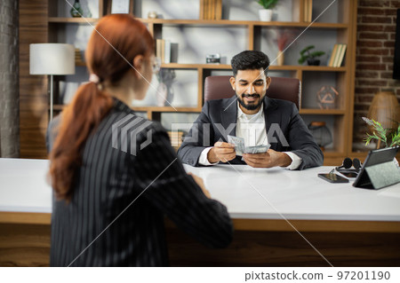 Businessman counting batch of hundred dollar bills sitting opposite his female business partner Businessman counting batch of hundred dollar bills sitting opposite his female business partner 97201190
