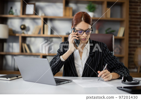 Portrait of pretty caucasian businesswoman working with laptop writes on document and calling phone Portrait of pretty caucasian businesswoman working with laptop writes on document and calling phone 97201430