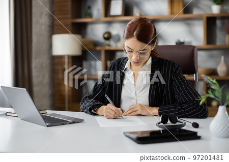Confident businesswoman wearing suit writing notes or financial report, sitting at desk with laptop Confident businesswoman wearing suit writing notes or financial report, sitting at desk with laptop 97201481