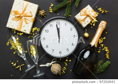 New year celebration concept with a bottle of champagne and two glasses toasting. Christmas gift box, alarm clock and fir tree branch on colored table. Top view Copy space 97202641