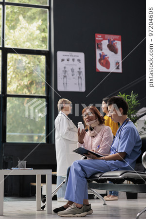 Medical assistant giving bottle pills to injured patient explaining medication treatment during medical appointment. Senior woman wearing cervical neck collar having consultation in hospital lobby Medical assistant giving bottle pills to injured patient explaining medication treatment during medical appointment. Senior woman wearing cervical neck collar having consultation in hospital lobby 97204608