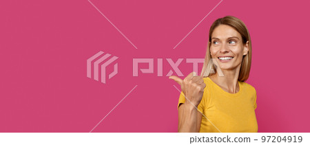 Smiling mature caucasian blond lady in casual point finger at copy space, isolated on pink background Smiling mature caucasian blond lady in casual point finger at copy space, isolated on pink background 97204919