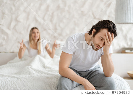 Angry woman shouting at her husband, having conflict while sitting on bed at home, focus on upset man Angry woman shouting at her husband, having conflict while sitting on bed at home, focus on upset man 97204923
