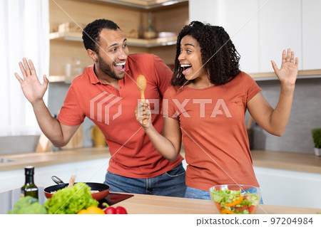 Glad young black man and woman in red t-shirts have fun, singing at imaginary microphone in kitchen interior Glad young black man and woman in red t-shirts have fun, singing at imaginary microphone in kitchen interior 97204984