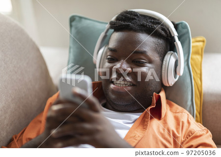 Black guy lying on couch with phone and headphones Black guy lying on couch with phone and headphones 97205036