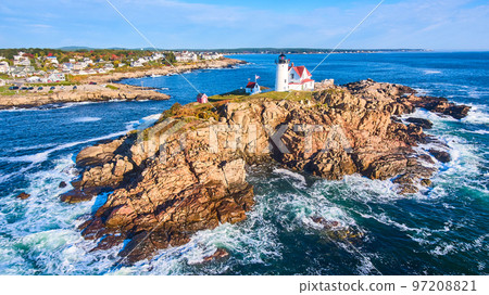 Waves crash over rocks aerial over Maine island with lighthouse and view of homes on mainland 97208821