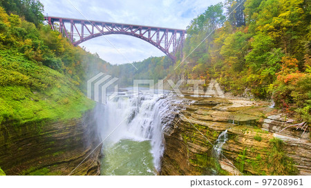 Steel maroon bridge over stunning raging waterfall cutting through layered cliffs in lush green forest Steel maroon bridge over stunning raging waterfall cutting through layered cliffs in lush green forest 97208961